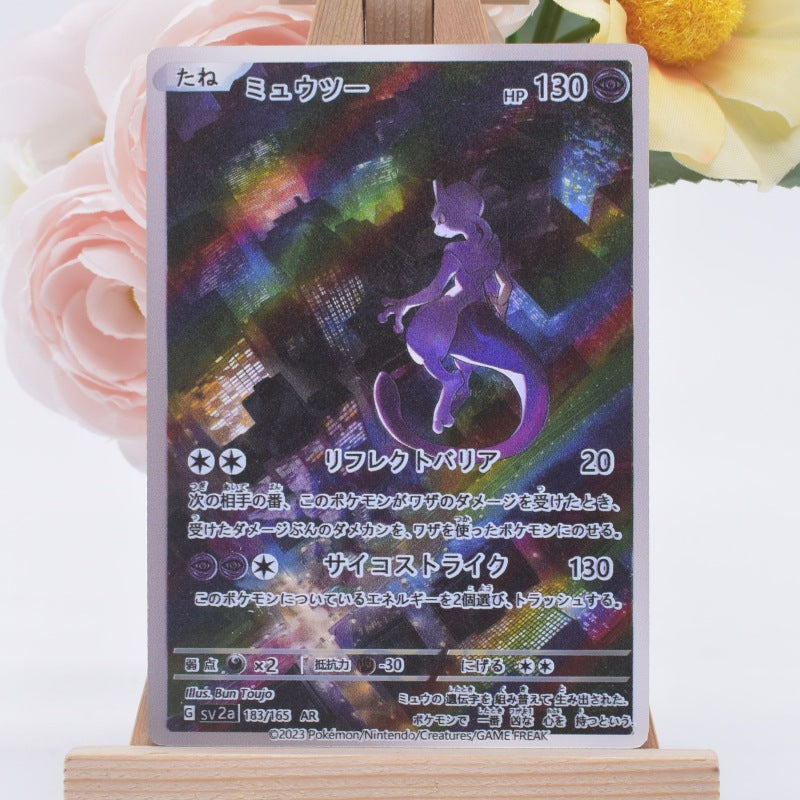 A holographic Pokémon trading card featuring Mew in a purple holographic finish, displayed on a wooden stand with pink and yellow flowers in the background.