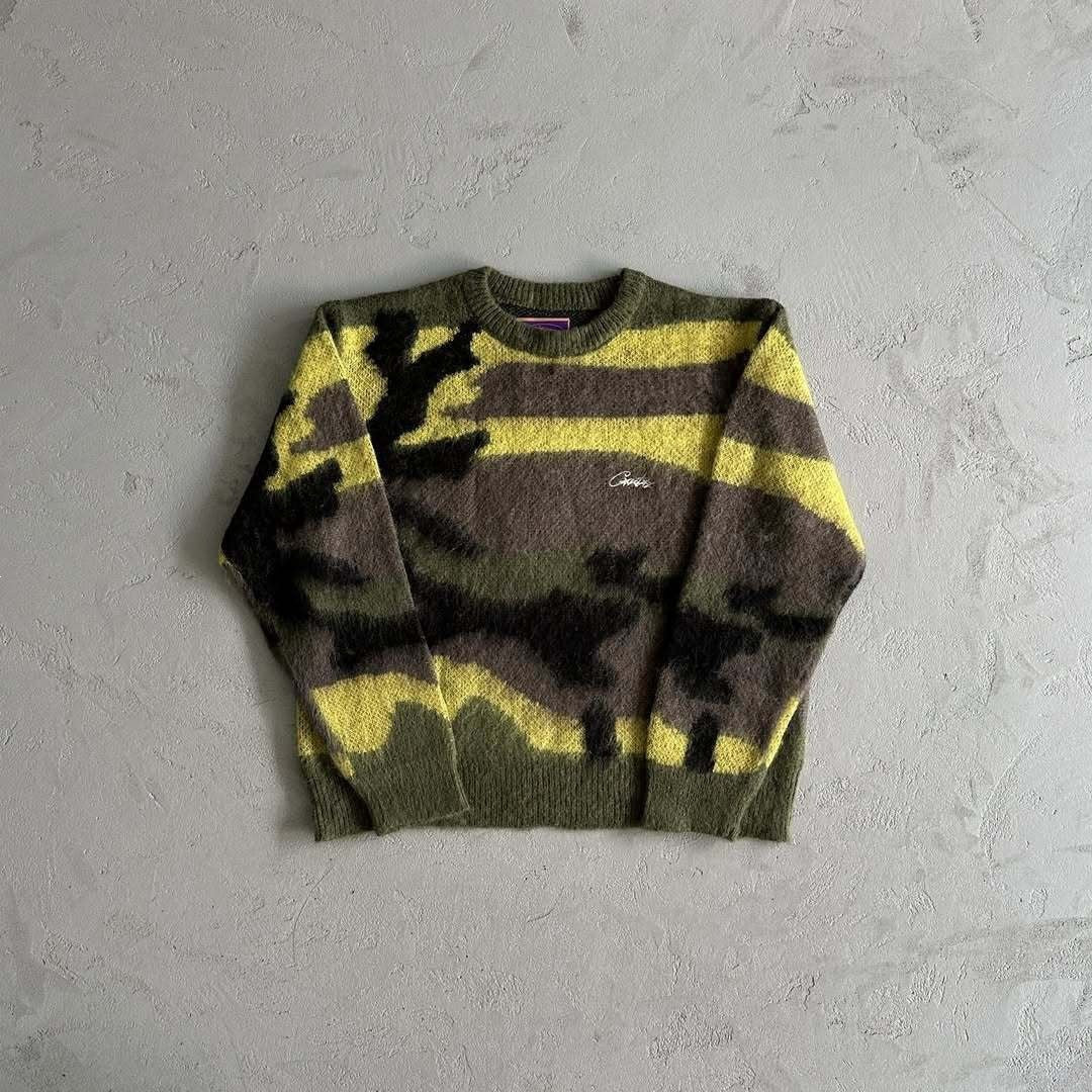 A fuzzy knit crewneck sweater with a camouflage pattern in olive green, brown, and yellow, laid flat on a textured light gray surface.