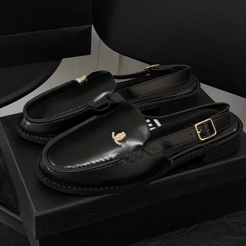 A pair of black leather slingback loafers with gold-tone decorative hardware, displayed on a black box against a dark studio background.