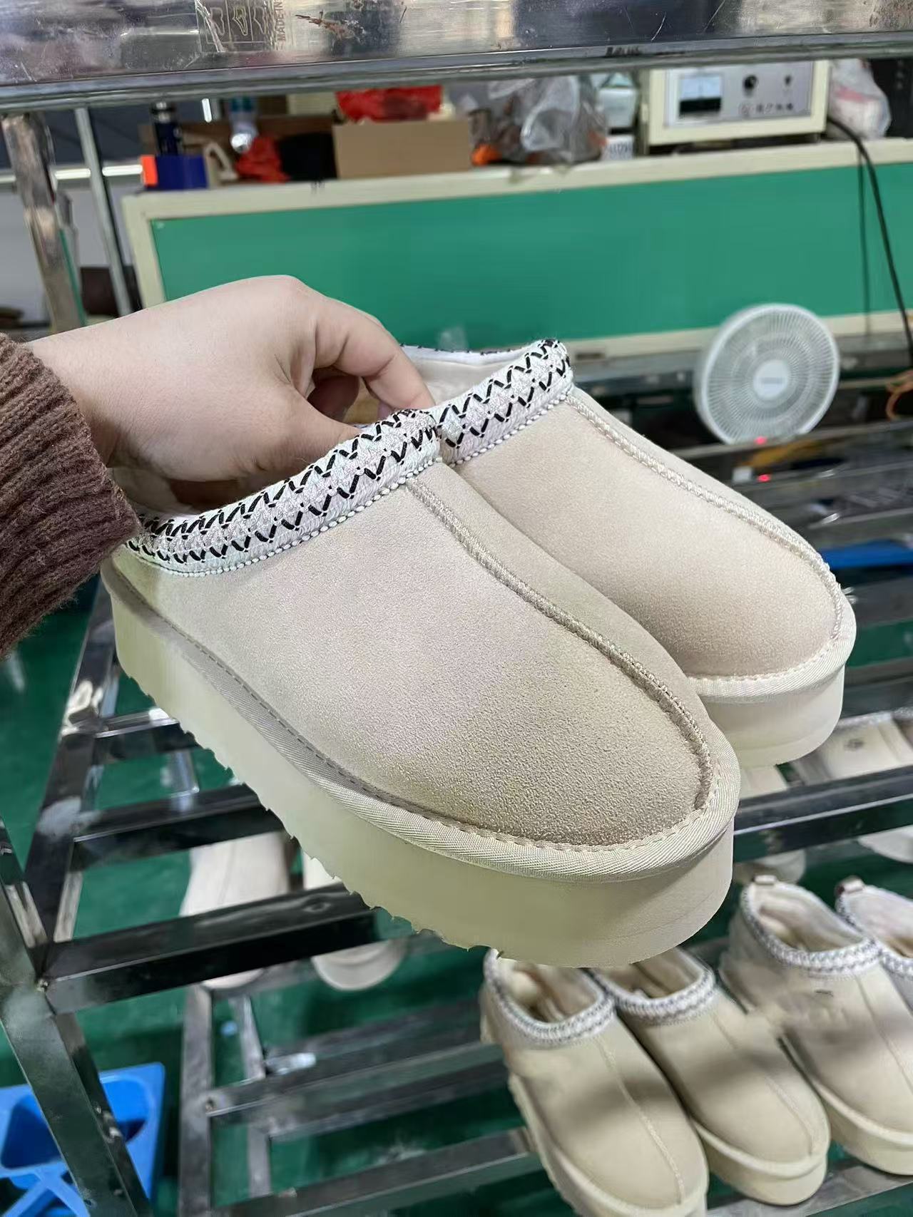 A pair of beige suede slip-on mule boots with a thick cream platform sole and a black-and-white zigzag trim along the collar, held by a hand in front of a metal shelving unit in a workshop.
