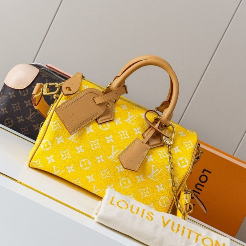 A yellow Louis Vuitton Speedy canvas handbag with a white monogram pattern, tan leather handles and tags, and gold-tone hardware, displayed on a shelf with a Louis Vuitton dust bag and branded box in the background.