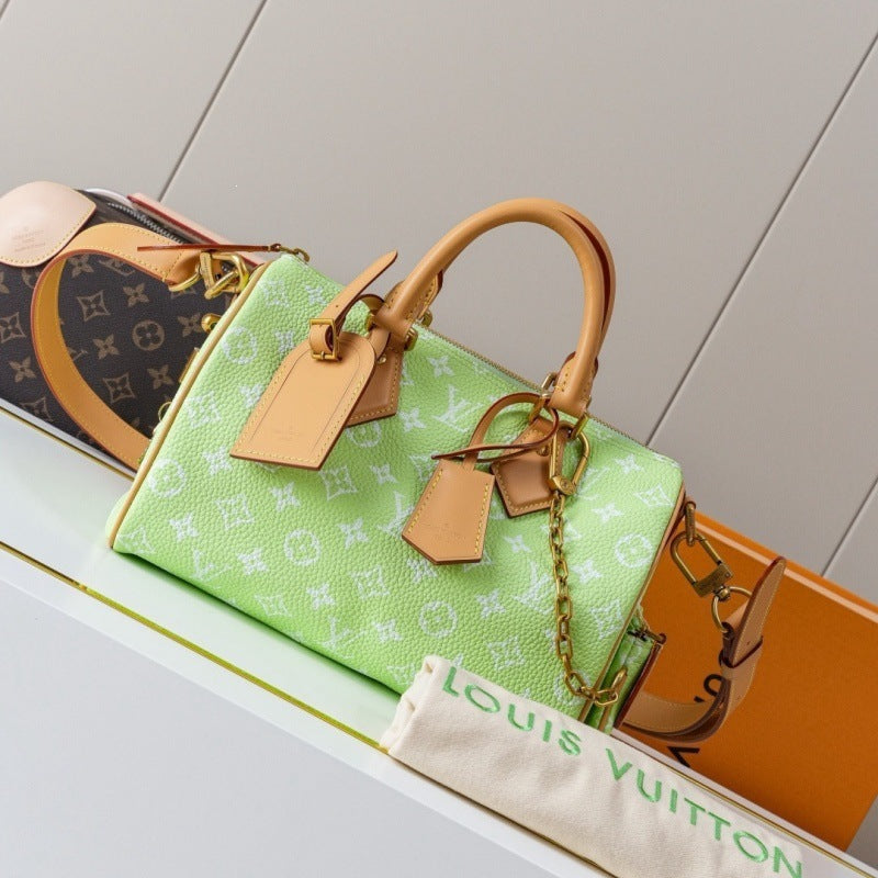 A light green Louis Vuitton monogram handbag with tan leather handles and tags, displayed on a white shelf next to other designer bags and a dust bag.