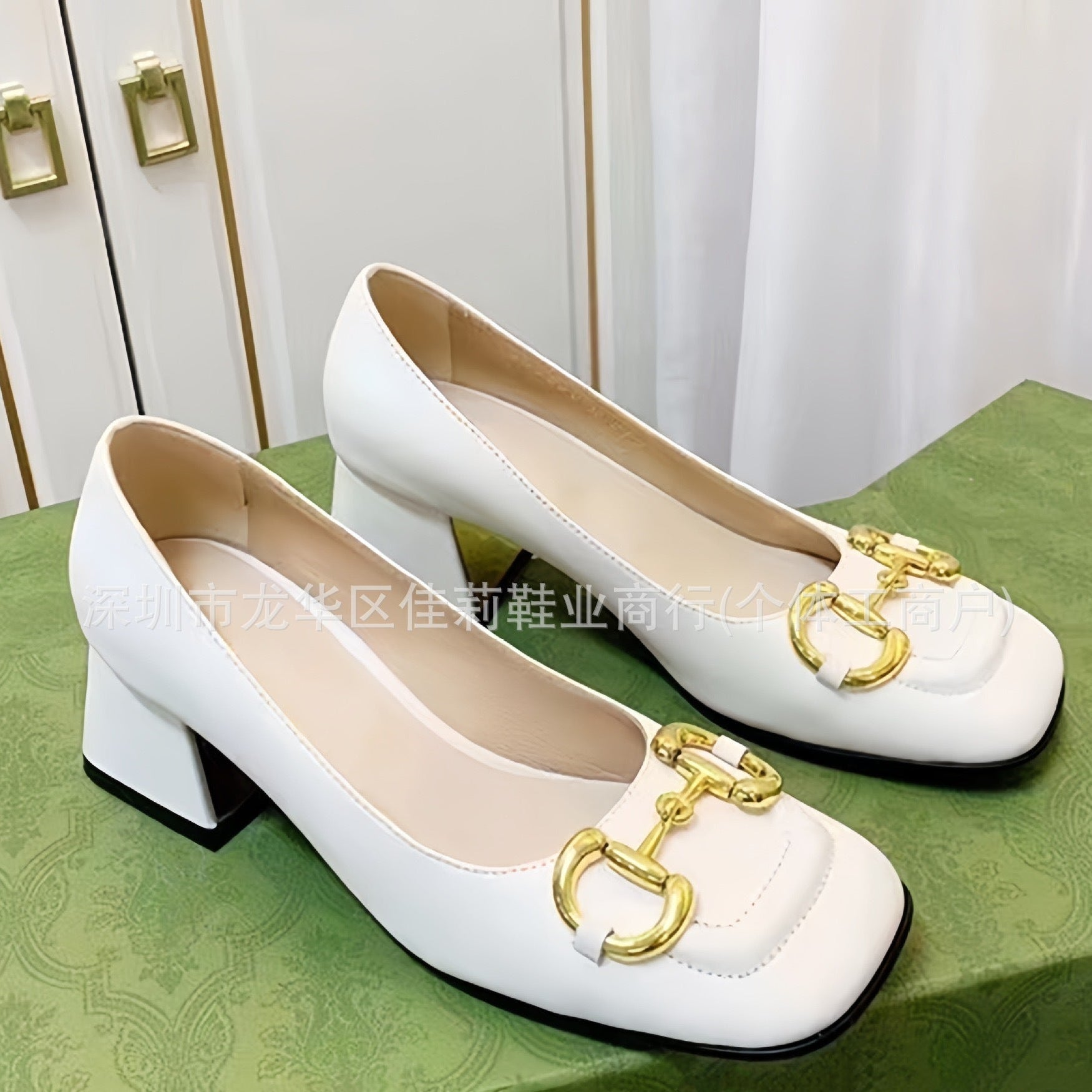 A pair of white leather block-heel pumps with a gold horsebit detail on the toe, displayed on a green patterned shoebox against a white curtain background.