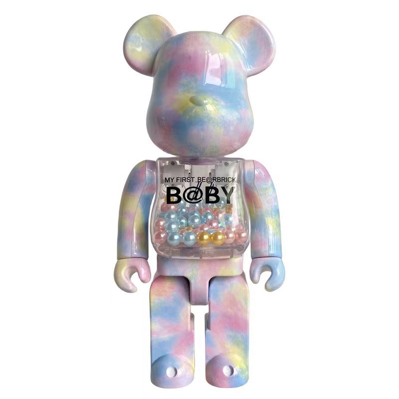 A pastel tie-dye Bearbrick toy figure with a transparent chest compartment filled with small pearls, shown against a plain white background. The chest has printed text reading 'MY FIRST BEARBRICK B@BY'.