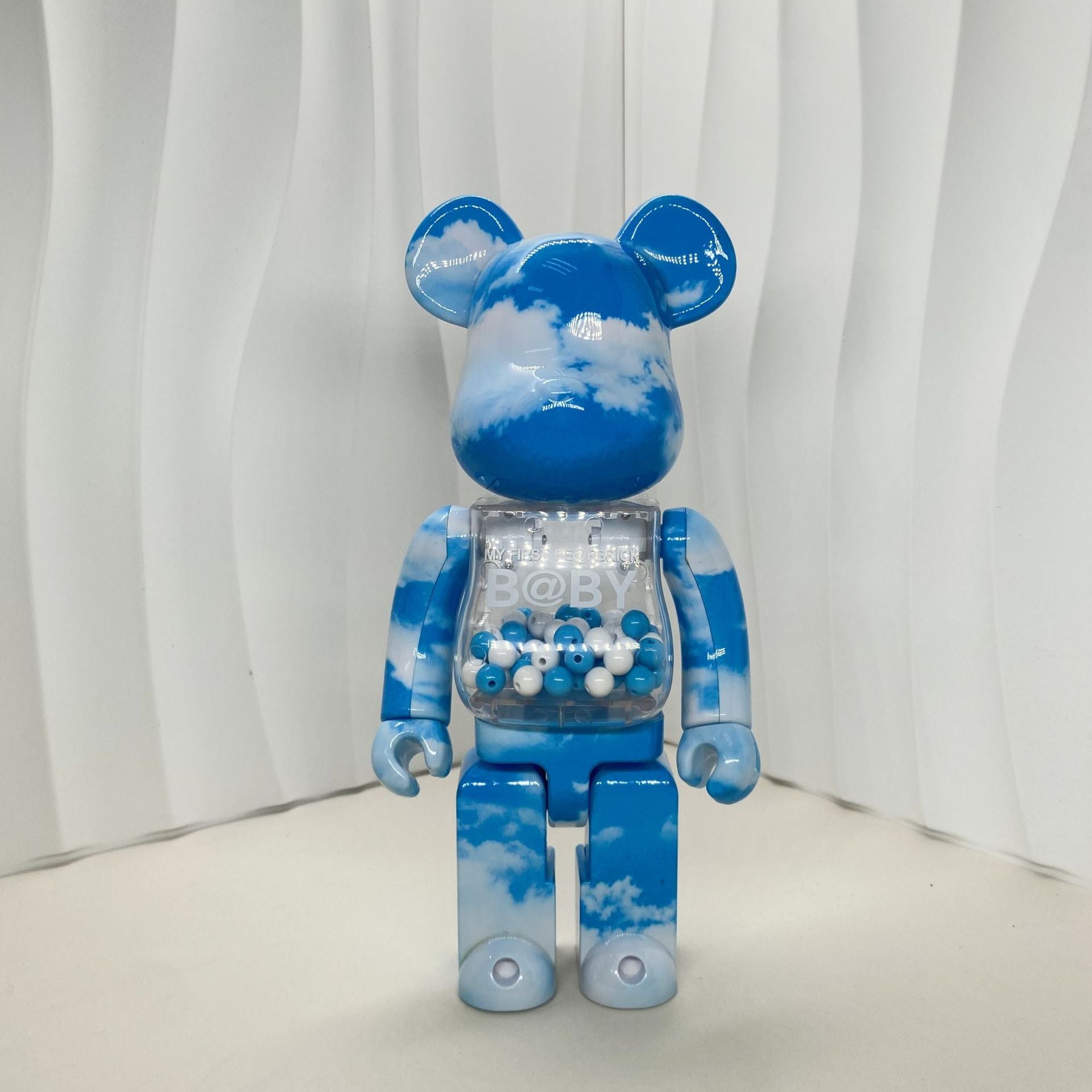A blue bear-shaped collectible figure with a sky-and-cloud print, standing against a white wavy backdrop. The torso has a transparent chest compartment filled with small blue and white beads.