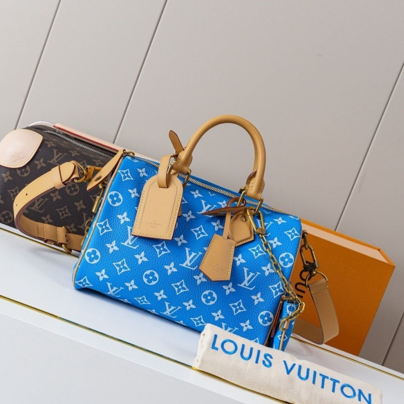 A Louis Vuitton blue monogram Speedy handbag with tan leather handles and a gold chain strap, displayed on a white shelf alongside other Louis Vuitton items and an orange box.