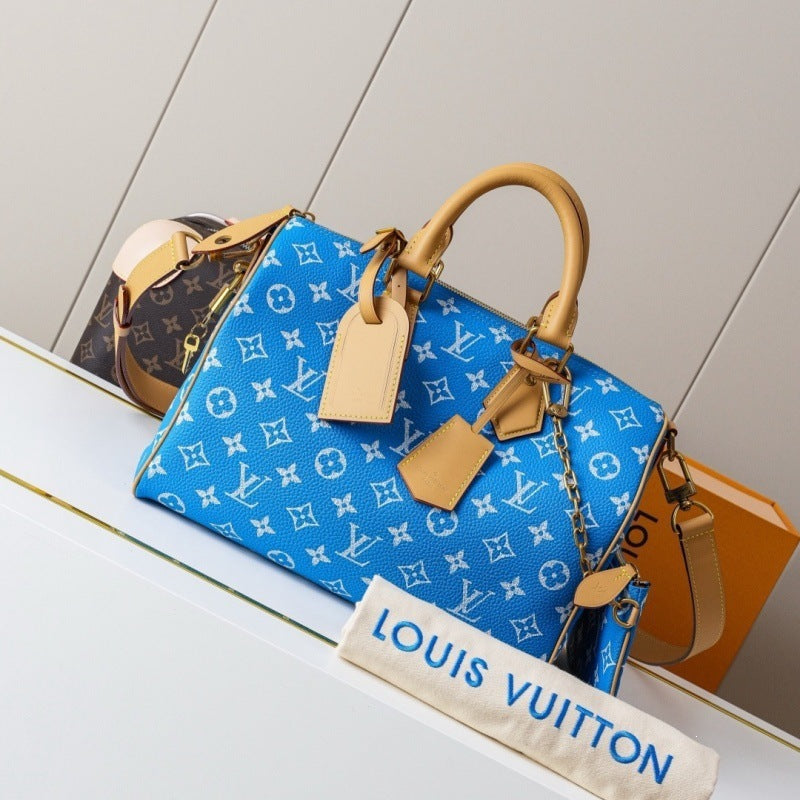 A blue Louis Vuitton monogram handbag with tan leather handles and trim, displayed on a white shelf with a Louis Vuitton dust bag in front and part of an orange box behind it.