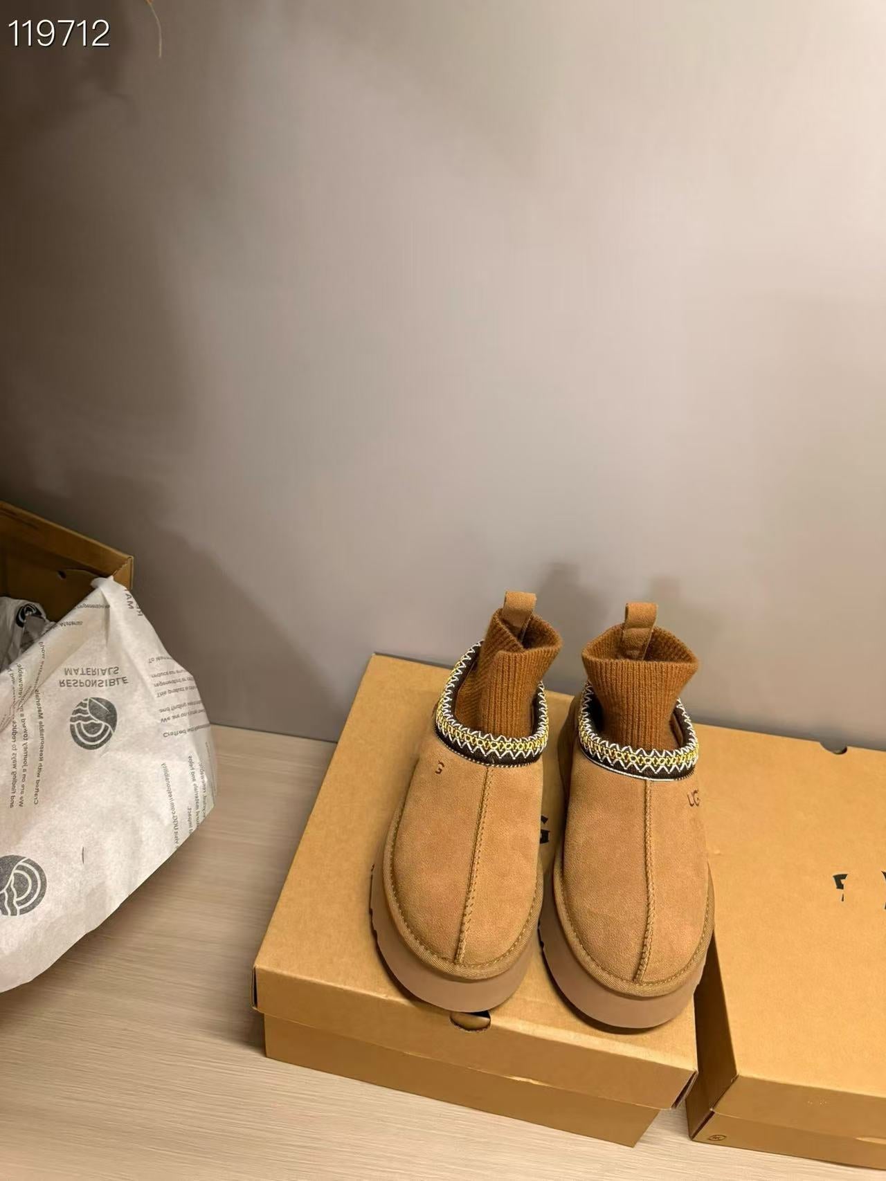 A pair of tan suede ankle boots with knit socks inside, resting on brown cardboard shoe boxes on a light wood floor against a gray wall.
