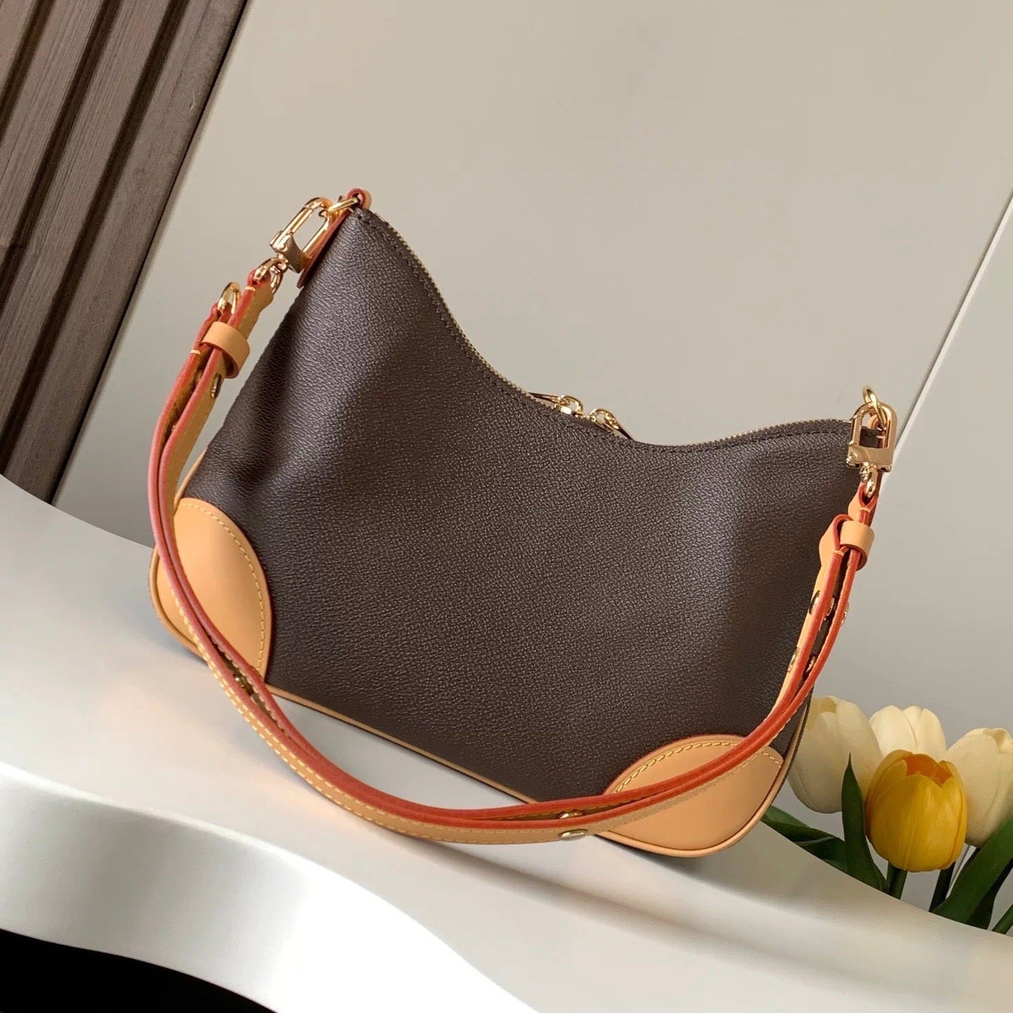 Brown handbag with orange strap on a white surface with flowers in the background