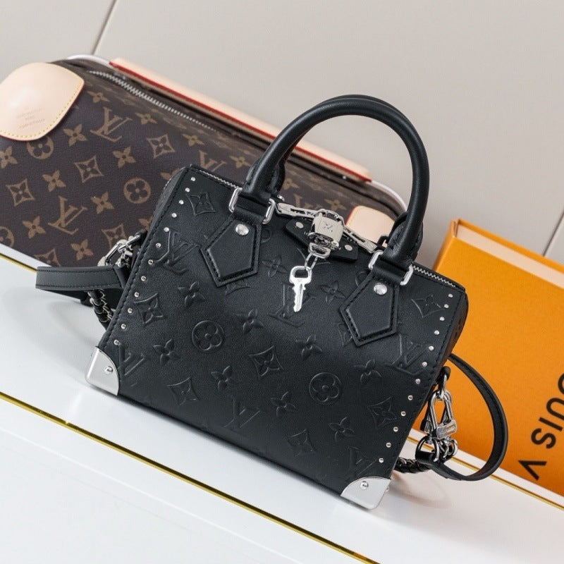 A black embossed leather handbag with silver-tone hardware and a key detail, styled on a white surface next to a Louis Vuitton monogram bag and an orange box.