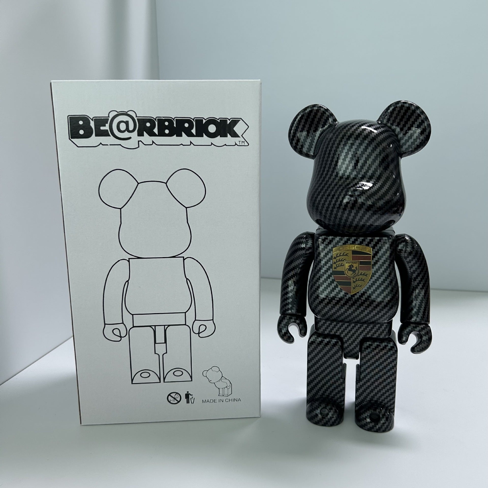 A Bearbrick collectible figure with a black carbon fiber pattern stands next to its white packaging box, which features the BE@RBRIK logo and a simple line drawing of the bear figure.