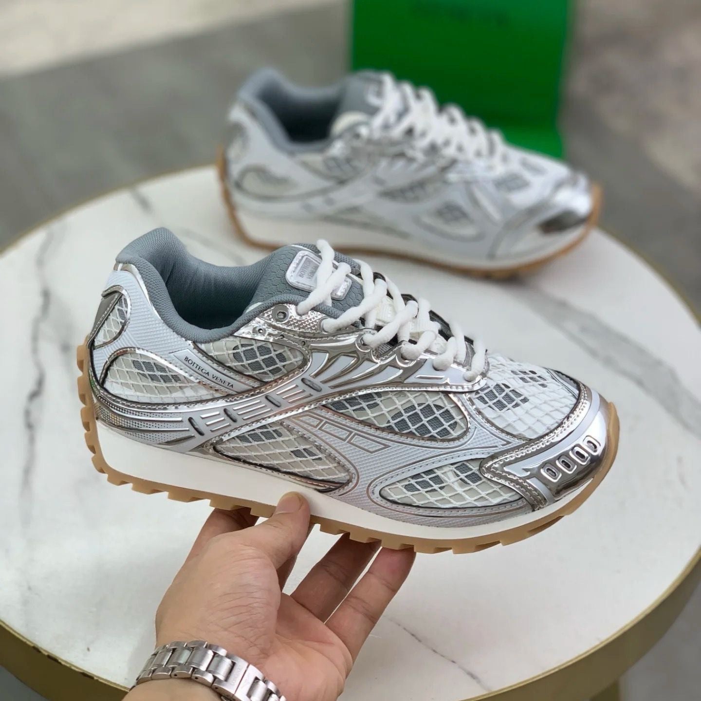 Silver sneaker with brown sole held by a hand on a marble surface