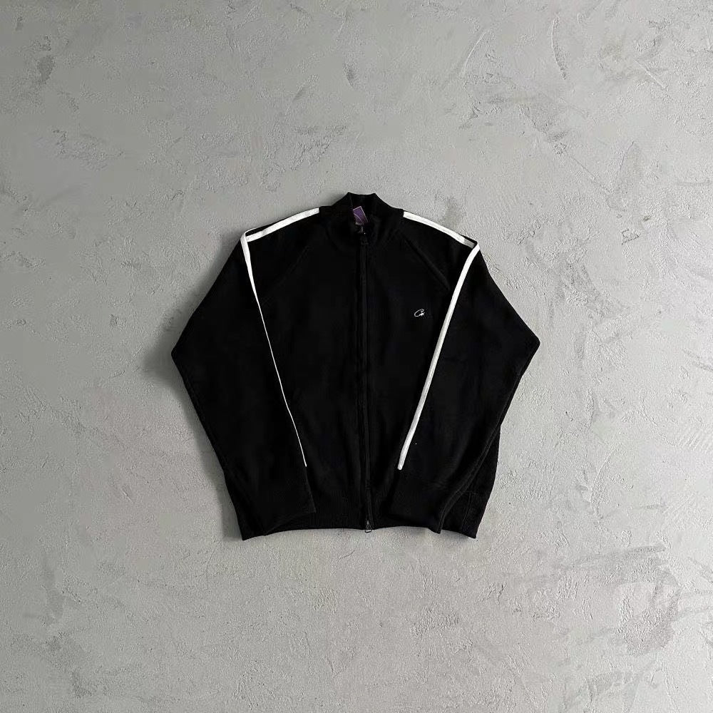 A black zip-up track jacket with white shoulder stripes, laid flat on a textured gray concrete background.