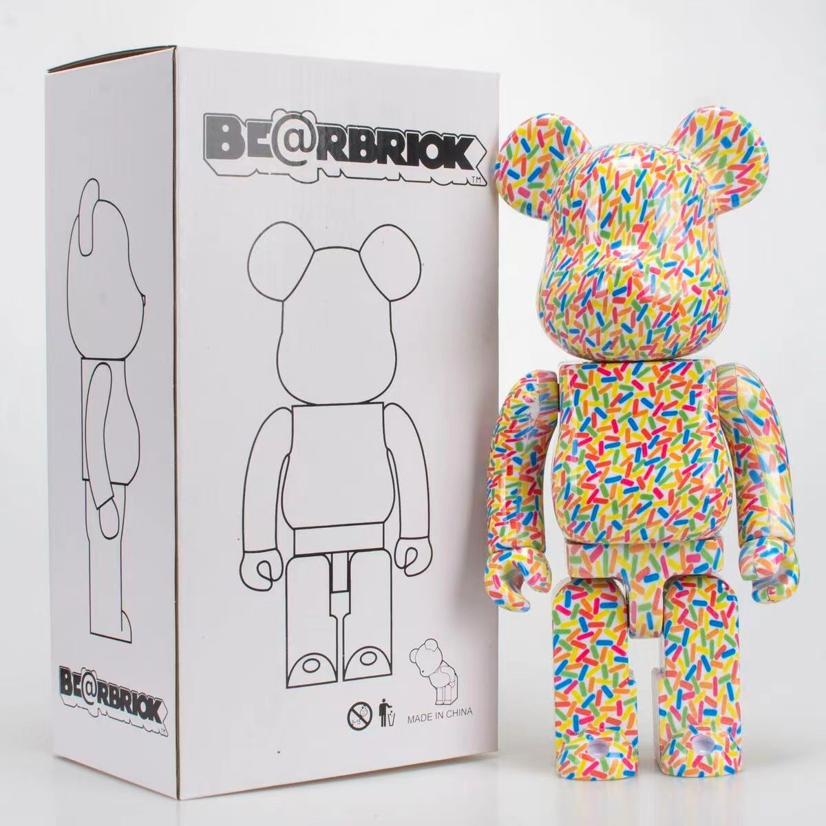 A Bearbrick collectible figure with a colorful sprinkle-patterned vinyl wrap stands next to its white cardboard packaging box, which features the BE@RBRICK logo and a black line drawing of the figure.