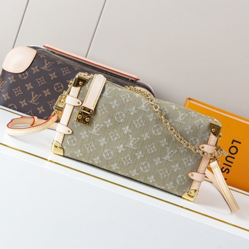 A Louis Vuitton monogram canvas shoulder bag in a muted sage green, styled on a white surface next to a Louis Vuitton monogram pouch and an orange Louis Vuitton box.