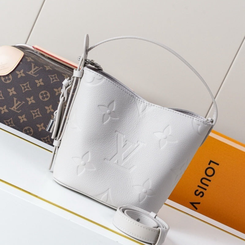 A light gray Louis Vuitton monogram-free leather bucket bag with a top handle, shown alongside a brown Louis Vuitton monogram pouch and an orange Louis Vuitton box on a white surface.