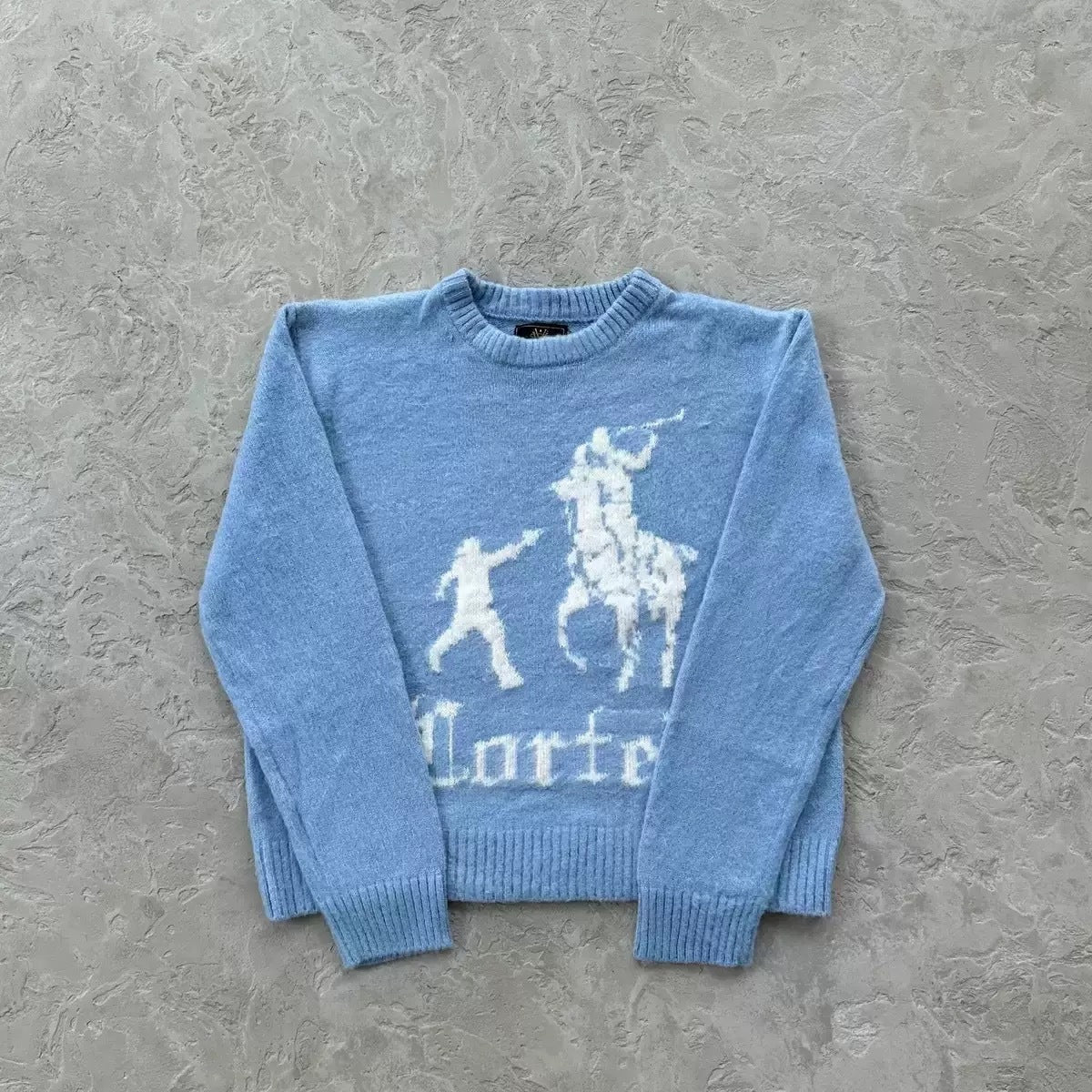 A light blue knit sweater with a white knitted graphic of a polo player and horse, and the word 'Porter' below, laid flat on a textured gray surface.