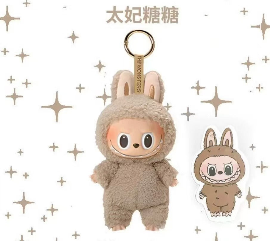 A plush keychain charm shaped like a cartoon character in a fuzzy brown bunny suit, shown with a metal key ring and a small matching die-cut sticker on a white background with decorative star motifs.