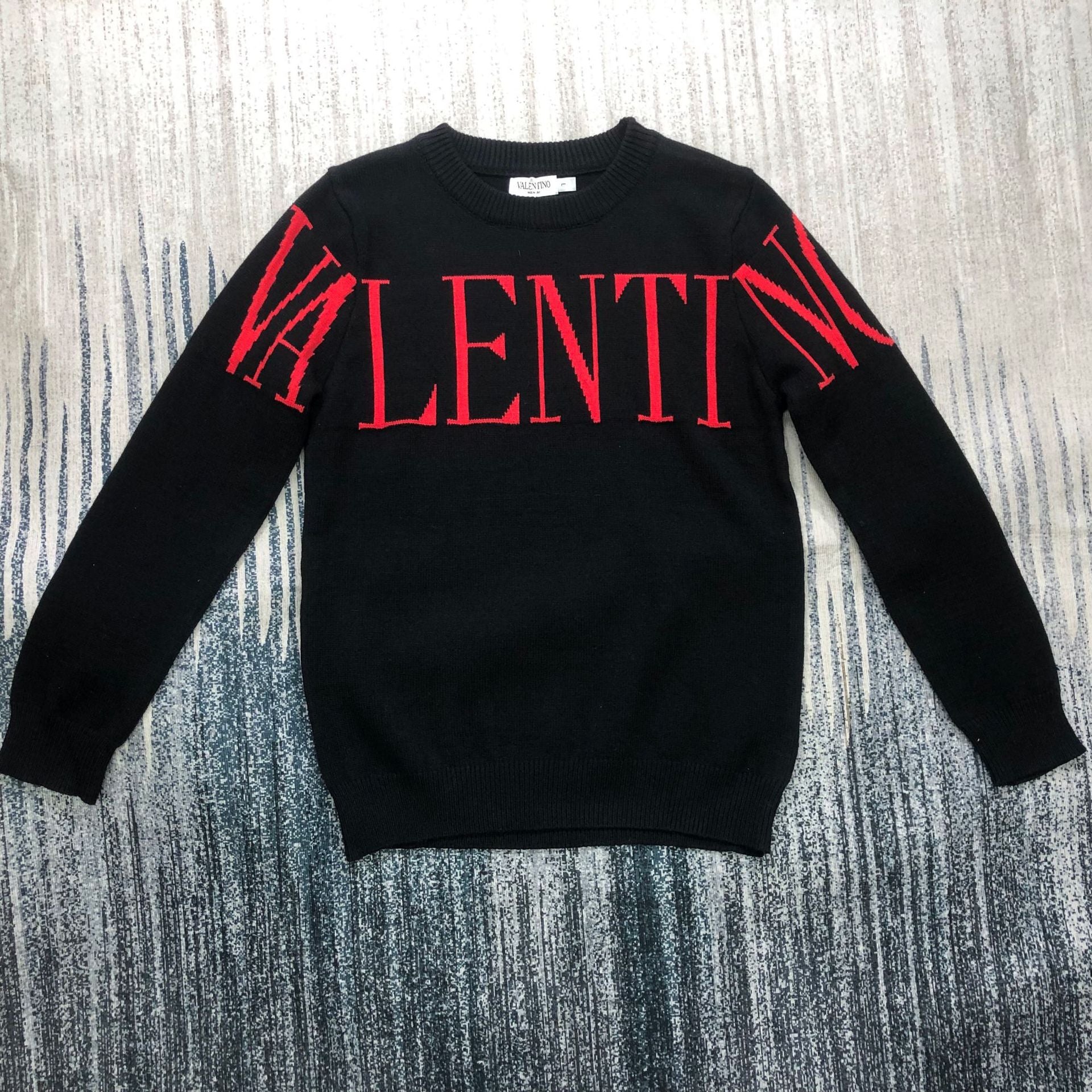 A black long-sleeve knit sweater with 'VALENTIN' written across the chest in red, laid flat on a gray and blue textured background.