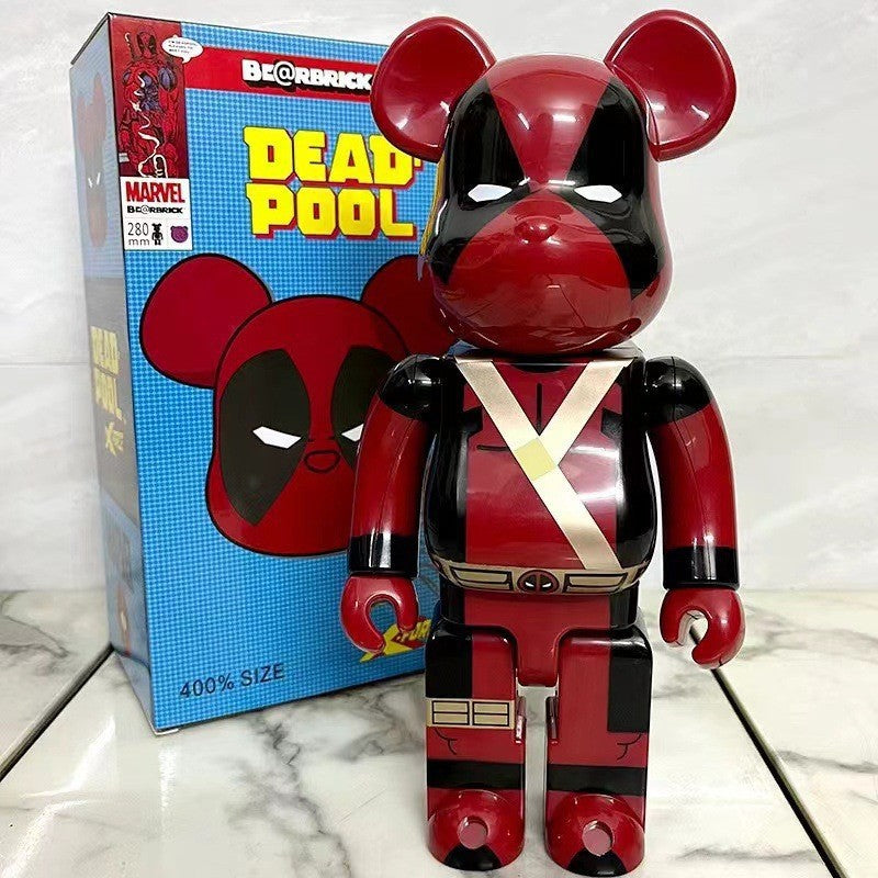 A red and black Bearbrick collectible figure of Deadpool standing in front of its blue and red packaging box on a light marble surface.