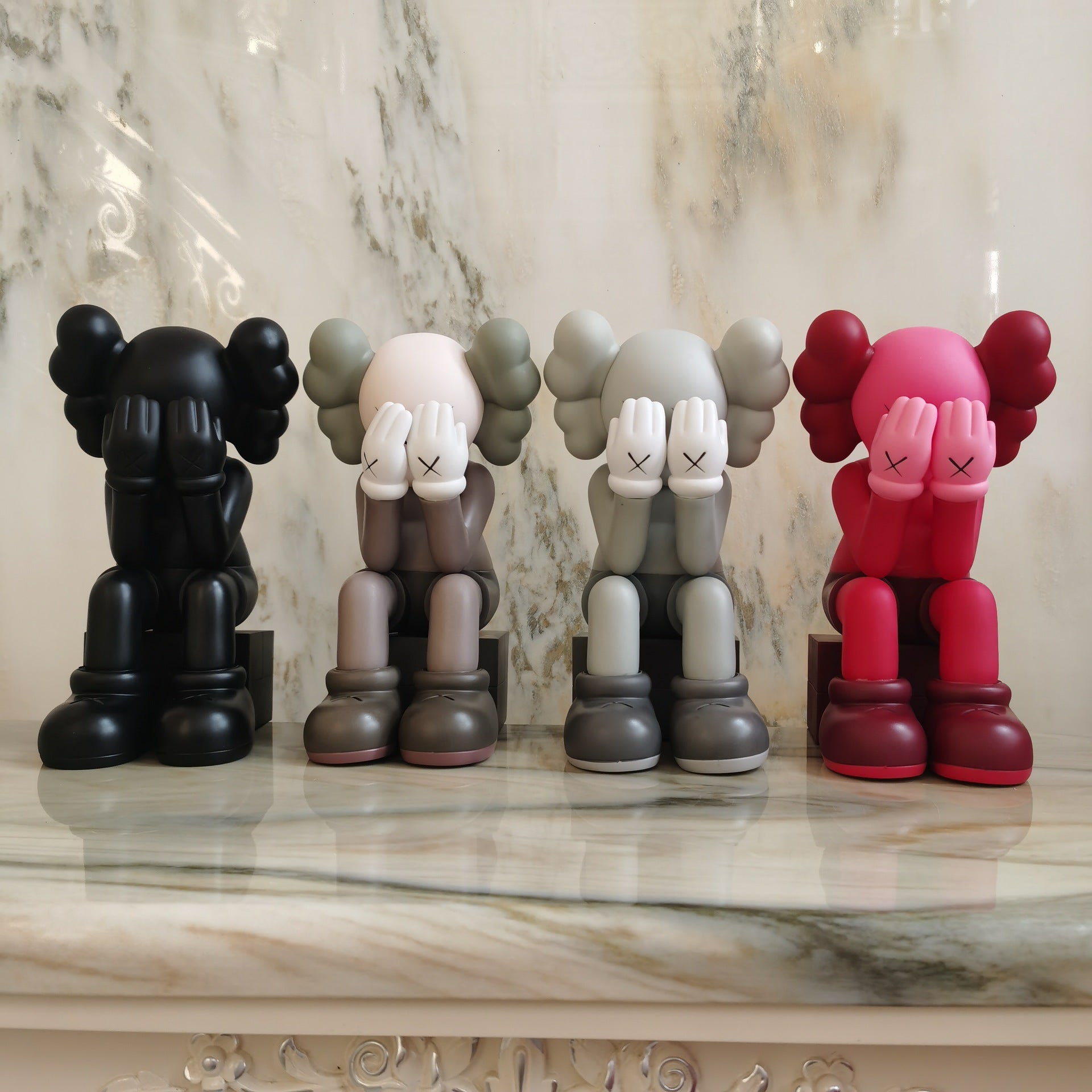 Four KAWS-style seated figurines in different colorways are lined up on a polished marble surface against a marble wall. The figures have the signature blindfolded pose with hands over eyes, shown in black, off-white/taupe, light gray, and bright pink/red.
