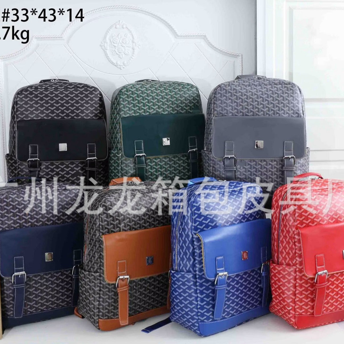 Six patterned backpacks in different colors are arranged against a white paneled wall. The backpacks include gray, black, green, blue, brown, and red, with a repeating monogram-style pattern and flap front pockets.