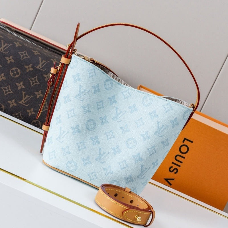 A Louis Vuitton handbag in light blue monogram canvas with brown leather trim and handle, displayed next to an orange Louis Vuitton box and a brown monogram pouch on a white surface.