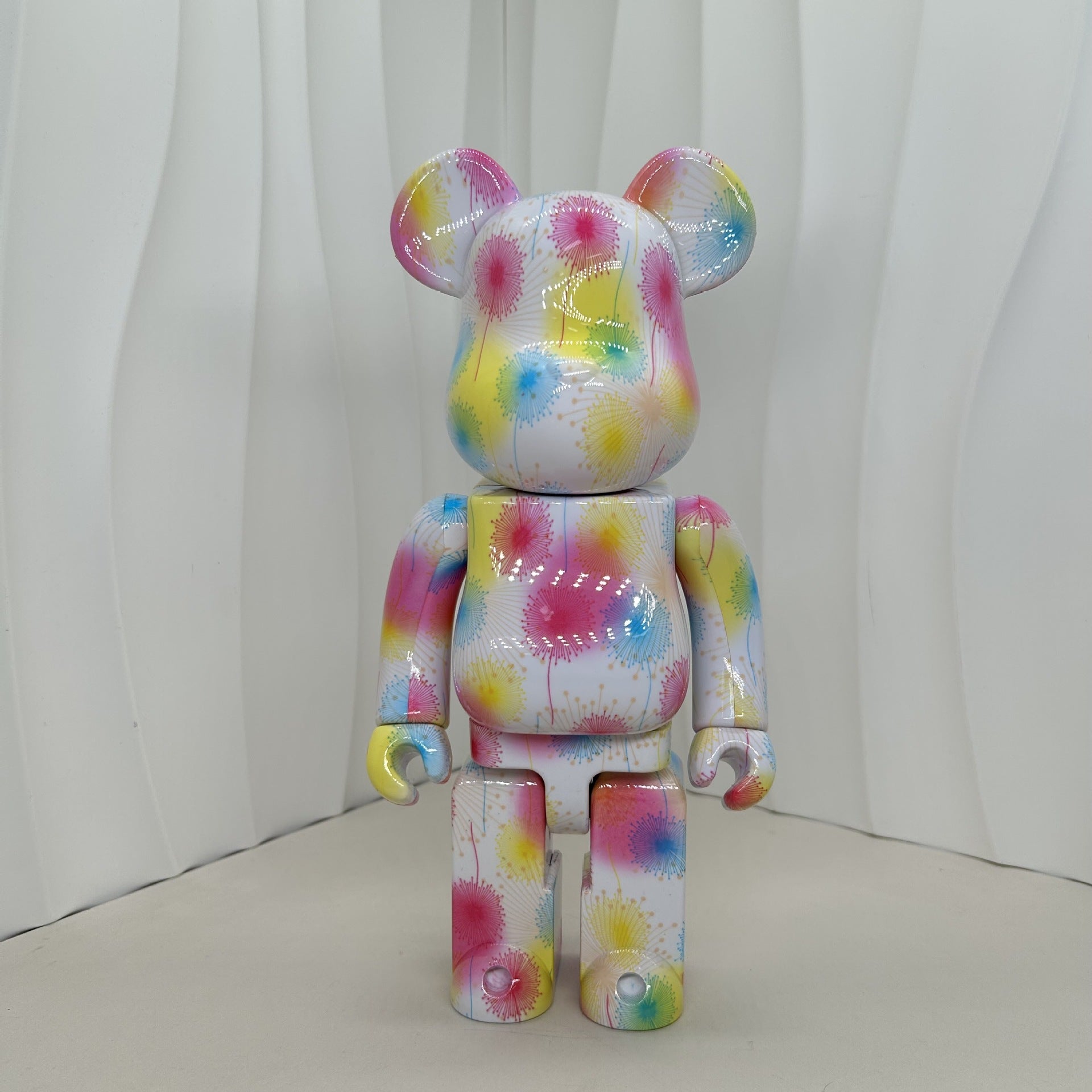 A bear-shaped collectible figure with a glossy white base covered in colorful paint splatter and drip patterns, standing against a white wavy backdrop.