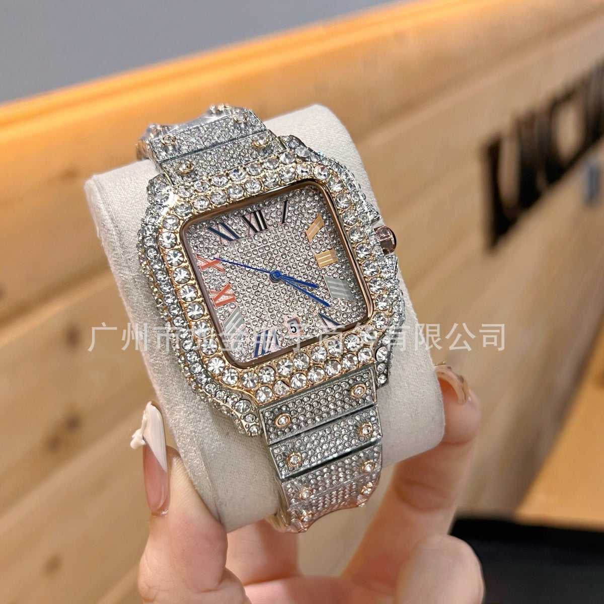 A close-up of a women's square analog watch encrusted with silver rhinestones, held on a cream-colored display stand by a hand. The watch features a textured silver dial with blue and orange accents, Roman numeral hour markers, and a matching rhinestone-embellished metal bracelet.
