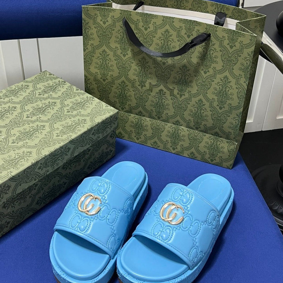 A pair of light blue slide sandals with embossed GG monogram patterns and a gold interlocking G logo on the strap, displayed with a matching green patterned box and shopping bag. The sandals are placed on a blue surface.