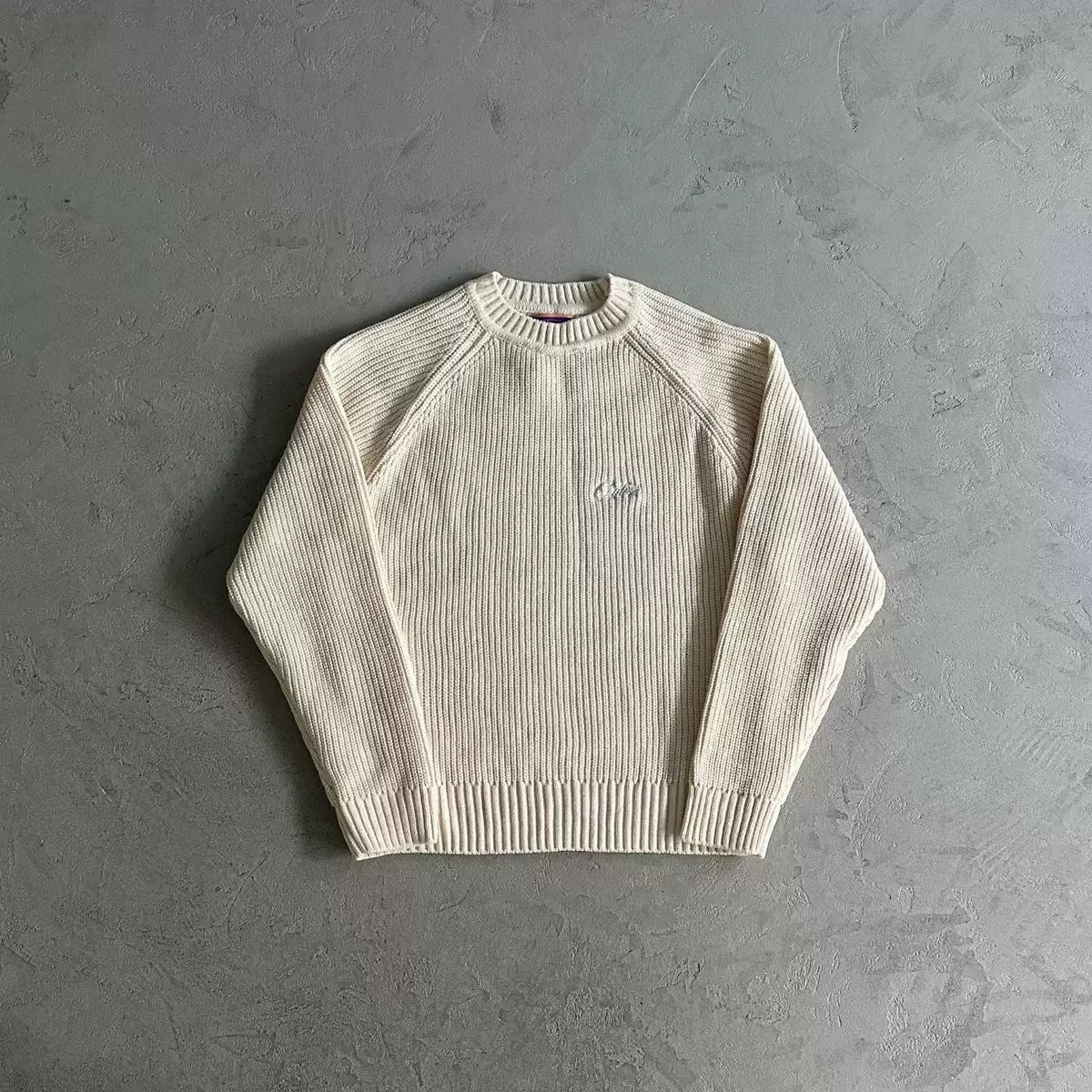 An off-white ribbed knit sweater with a small embroidered logo on the left chest, laid flat on a textured gray concrete surface.