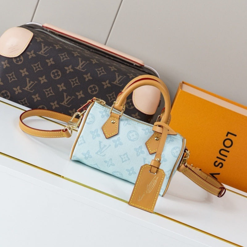 A light blue Louis Vuitton Speedy handbag with tan leather trim and handles, displayed next to a dark brown Louis Vuitton monogram pouch and an orange Louis Vuitton box on a white surface.
