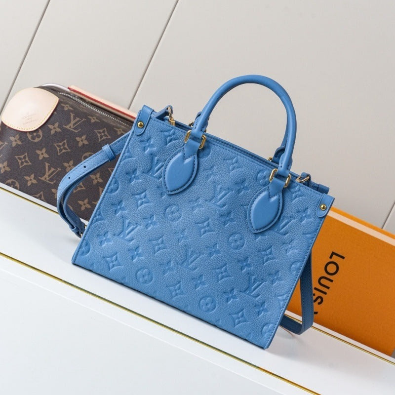 A blue Louis Vuitton monogram handbag with double top handles and a shoulder strap, styled on a white surface with a Louis Vuitton box and a brown monogram pouch in the background.