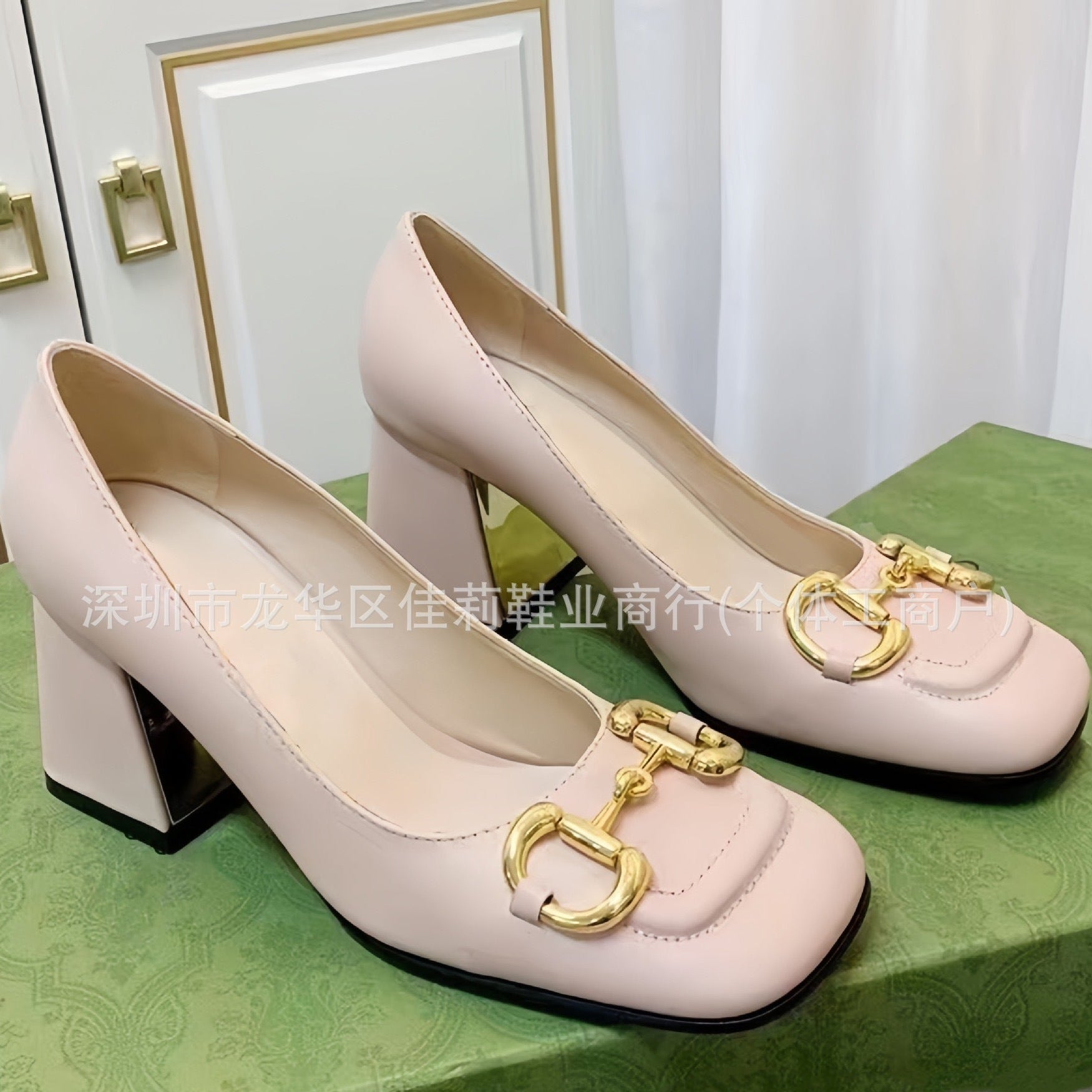 A pair of women's block-heel pumps in pale blush pink with gold horsebit hardware on the toes, displayed on a green patterned surface.
