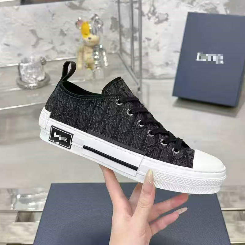 A hand holds a black low-top sneaker with an allover monogram pattern, a white rubber toe cap and sole, and a black heel patch. A matching dark blue shoebox is visible in the background on a marble-patterned surface.