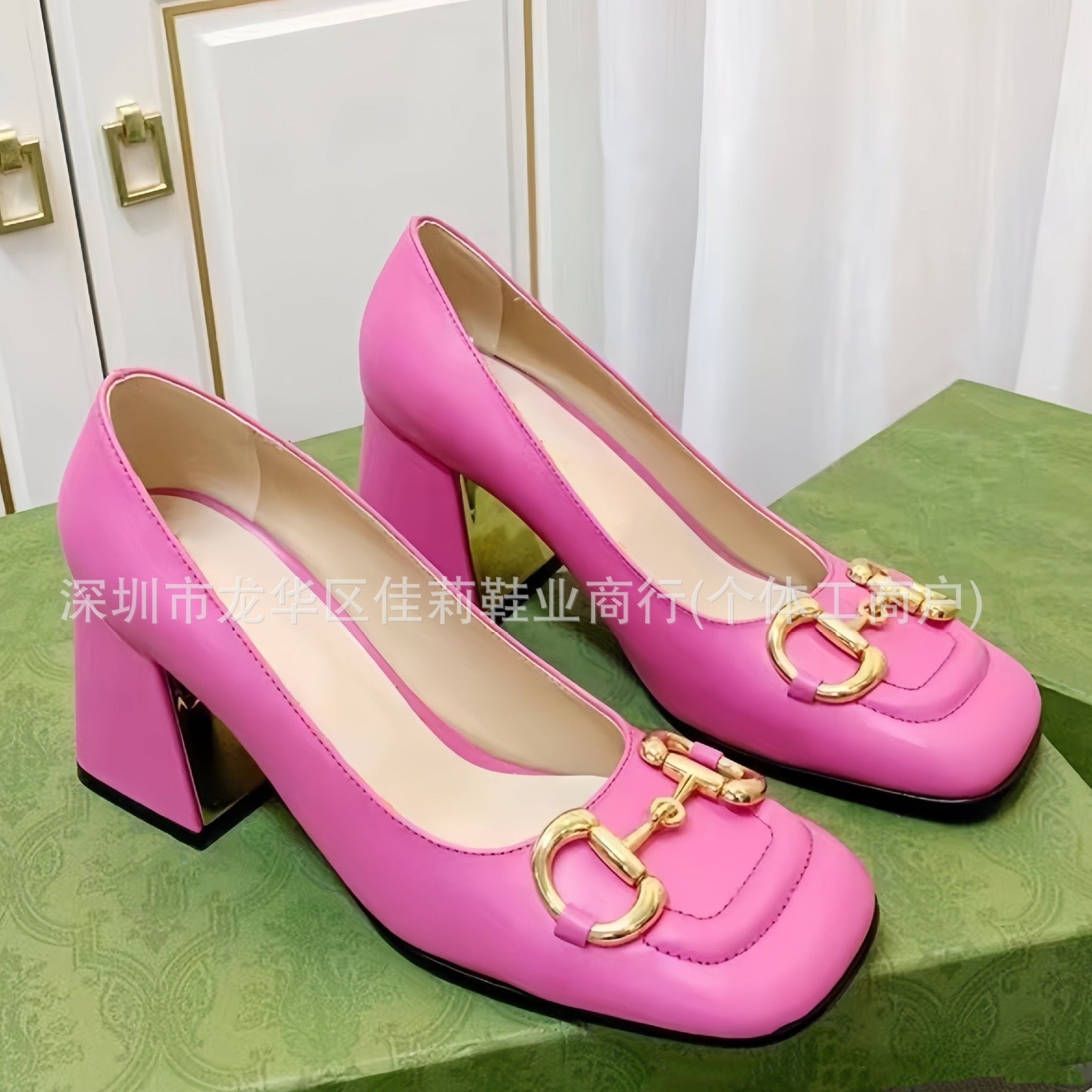 A pair of bright pink block-heel pumps with gold horsebit hardware on the toes, displayed on a green shoebox against a white curtain background.