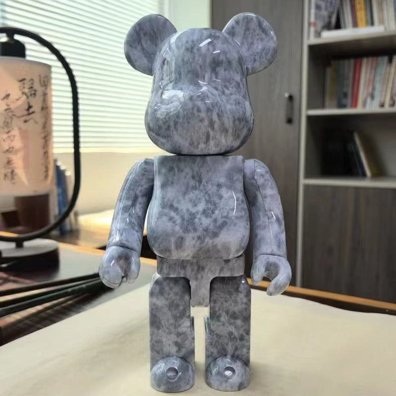 A bear-shaped collectible figure with a gray and white marbled pattern, standing on a tabletop in an indoor room with shelves and a lamp in the background.