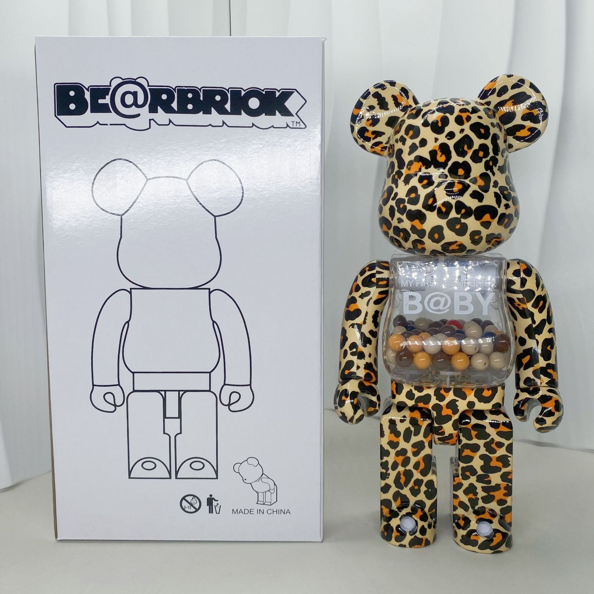 A leopard-print Bearbrick toy stands next to its white product box, which features a black line drawing of the bear figure. The bear has a clear plastic chest compartment filled with wooden beads, and 'BE@RBRIOK' and 'B@BY' branding are visible.