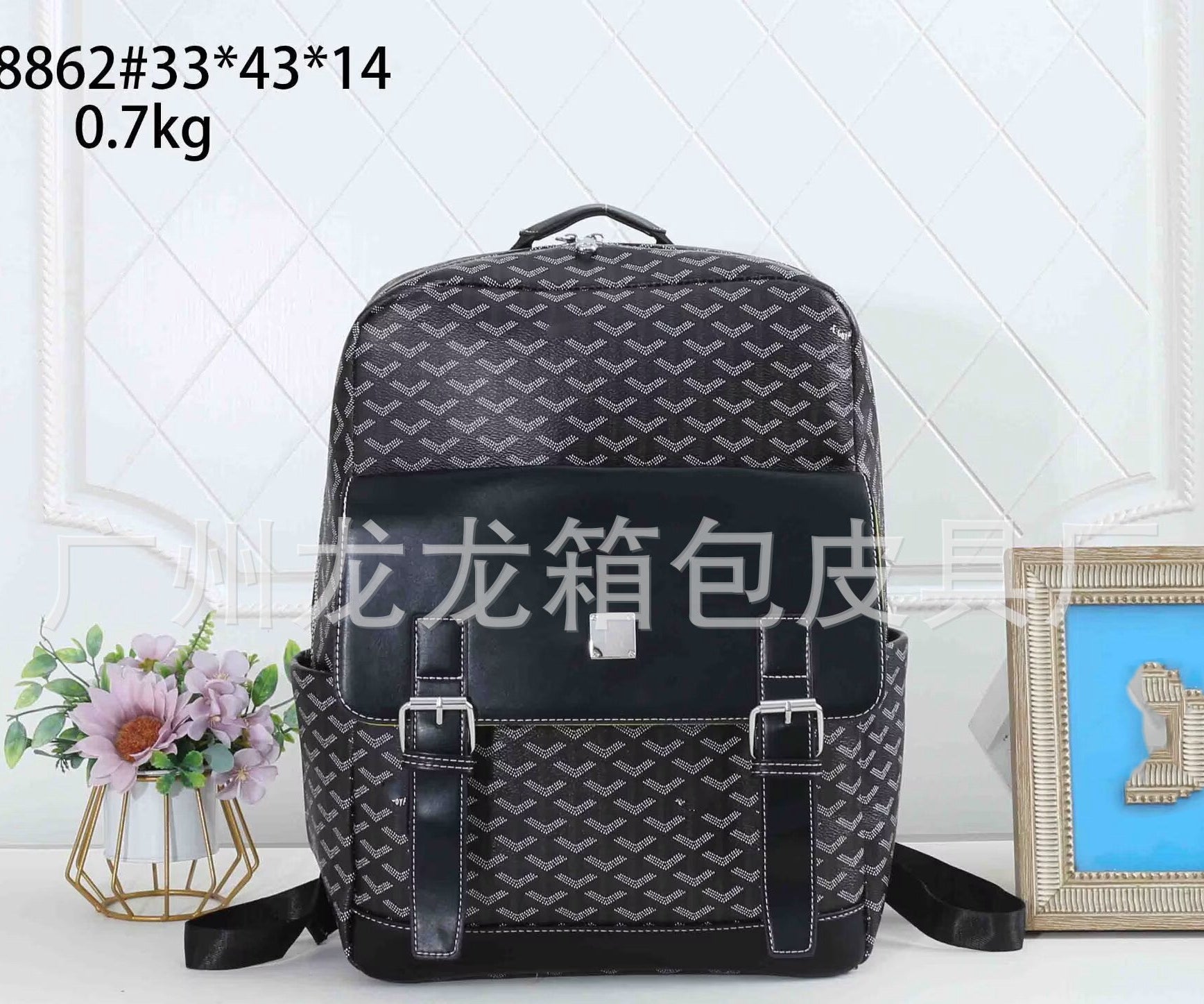 A black and gray patterned backpack with a large black front pocket and silver buckles, displayed on a white surface with decorative props including a small flower vase and a framed picture. Text overlays show product details.
