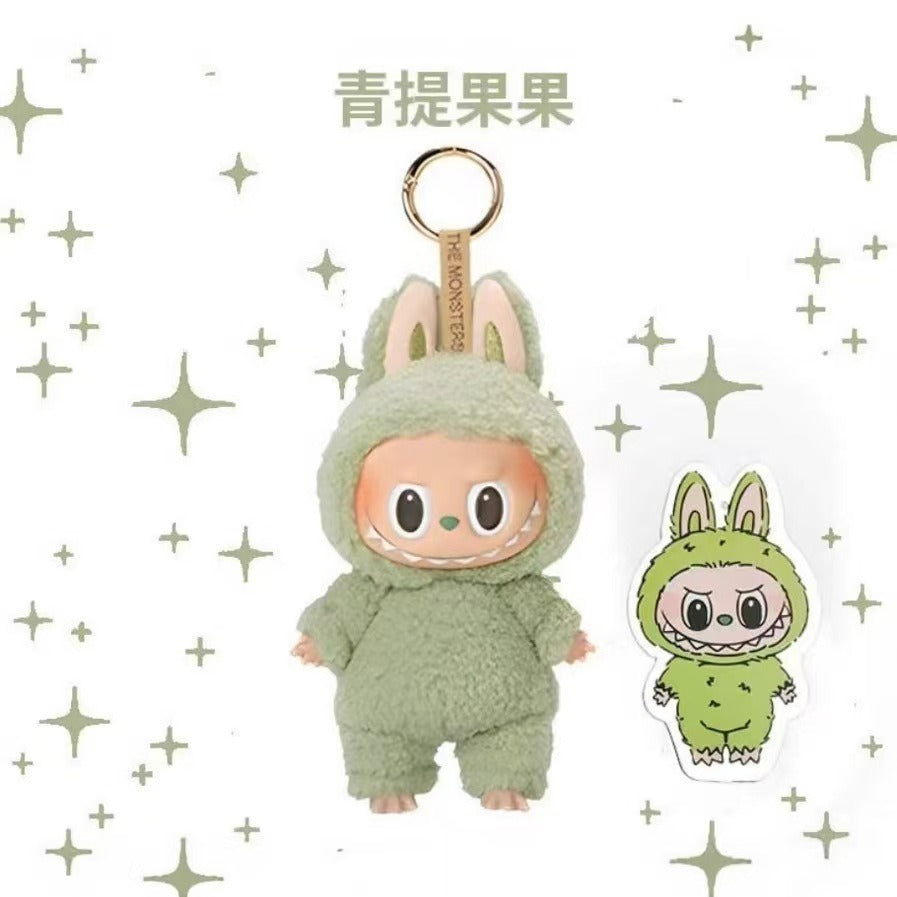 A plush character keychain in a fuzzy sage green bunny suit with a peach-toned face and large eyes, attached to a gold ring. A matching die-cut sticker is shown to the right on a white background with scattered star motifs and the Chinese text '青提果果' above the product.