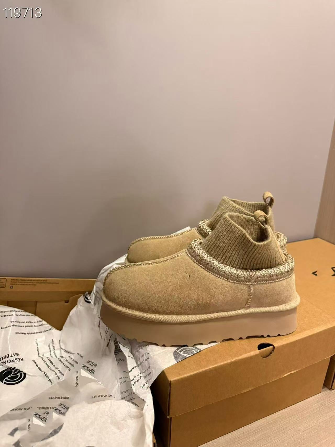 A pair of beige suede ankle boots with knit cuffs and chunky platform soles, resting on a cardboard shoebox with branded tissue paper.