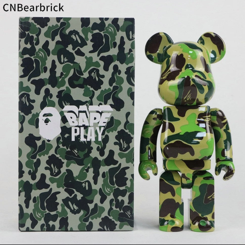 A green and black camouflage A Bathing Ape Bearbrick toy figure standing next to its matching camouflage packaging box with a white BAPE PLAY logo, on a light gray background.