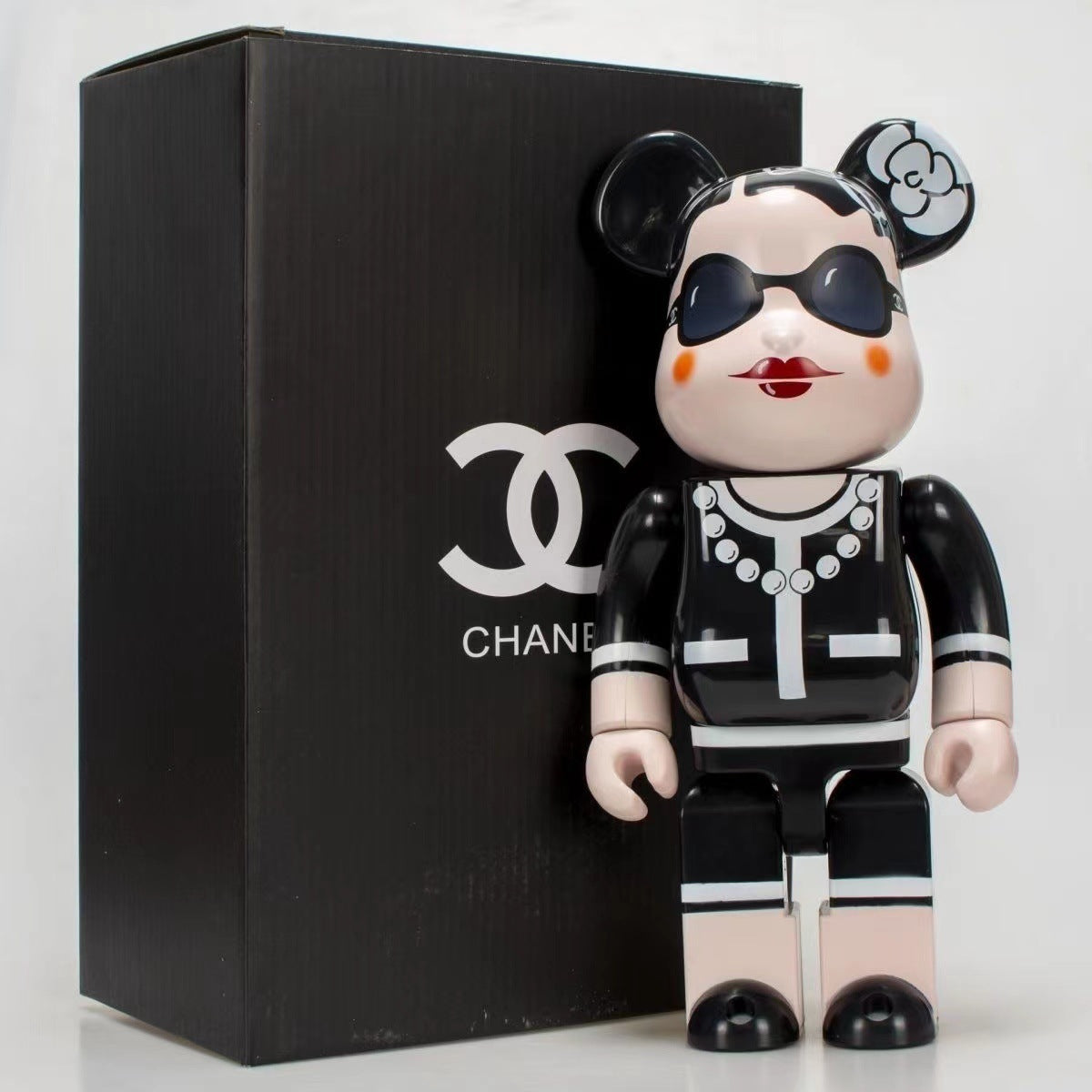 A black and white Chanel-themed bearbrick figure with sunglasses, a flower hair accessory, and red lips, standing in front of a black Chanel box.