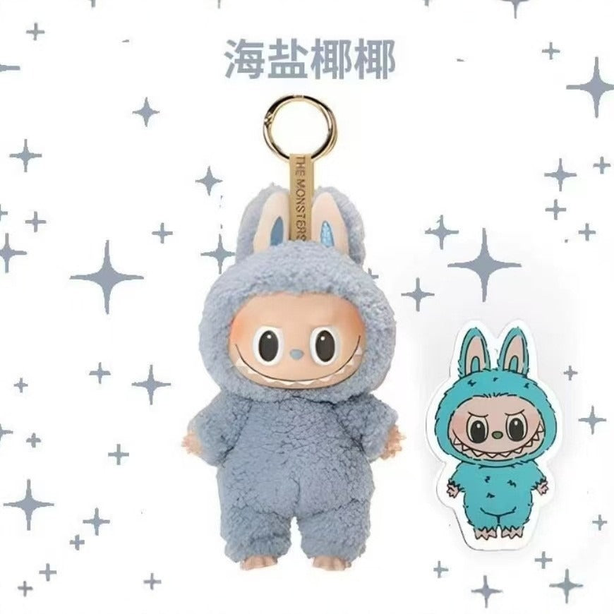 A plush character keychain or bag charm shown on a white background with scattered gray star graphics. The main subject is a fuzzy light blue bunny-like character with a beige face and a gold metal ring at the top.