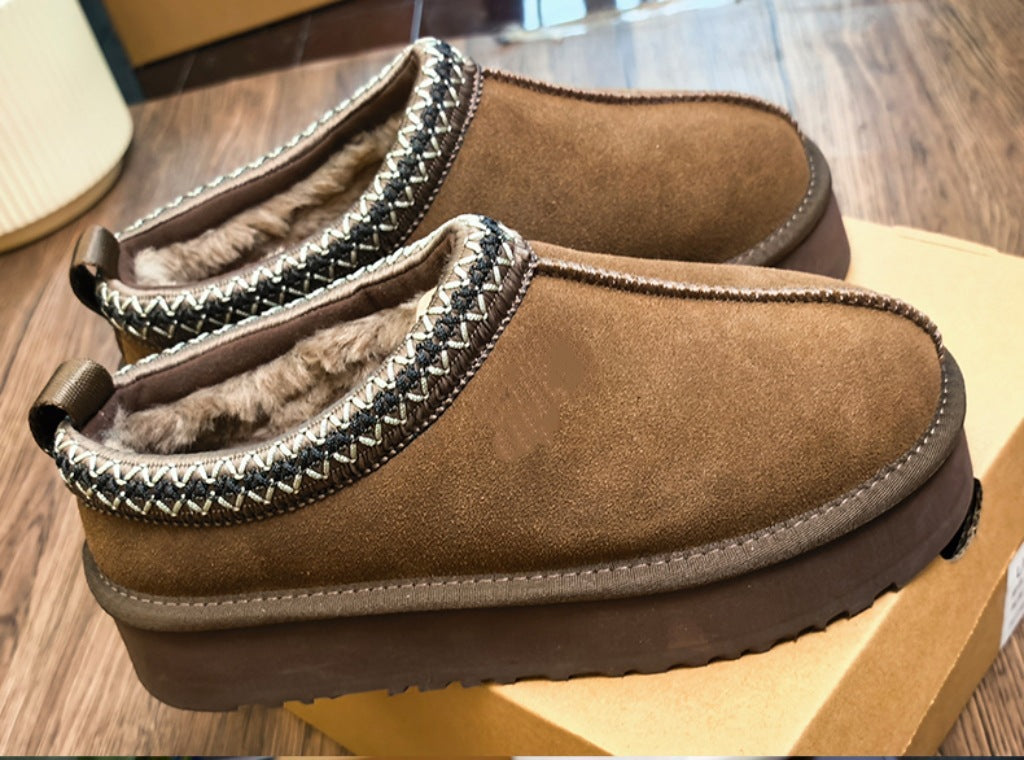 A pair of brown suede slip-on slippers with a soft faux-fur lining and a decorative black and beige geometric trim, resting on a cardboard shoebox over a wooden floor.