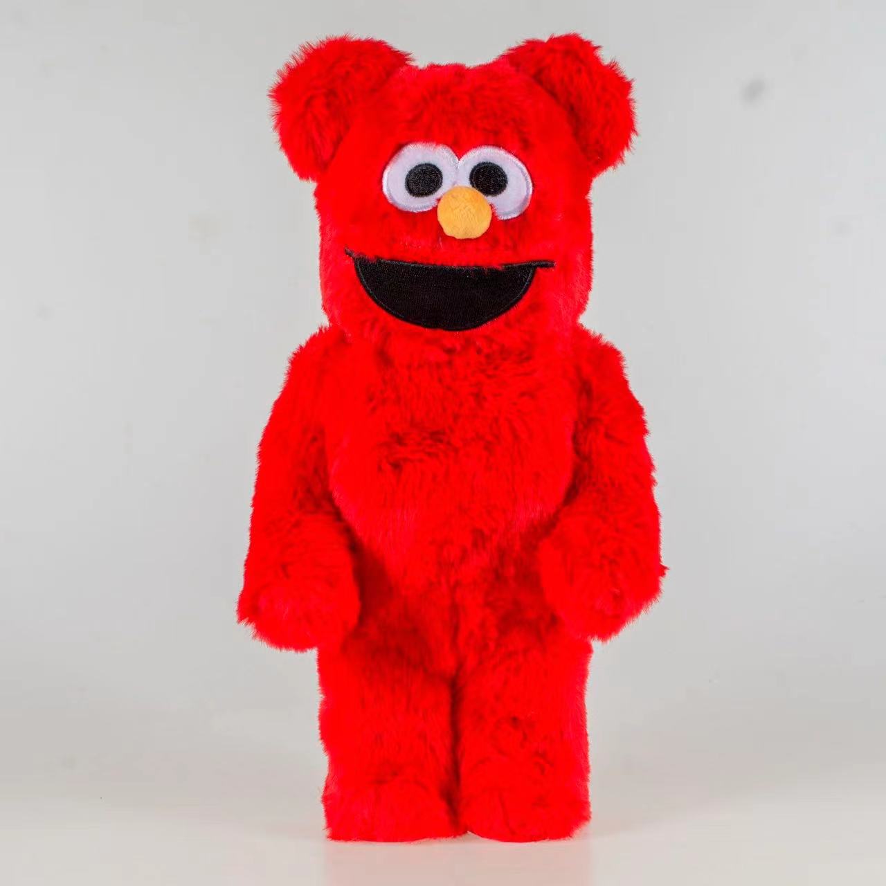 A red plush bear toy with large white eyes, a yellow nose, and a black mouth, standing against a plain light gray background.