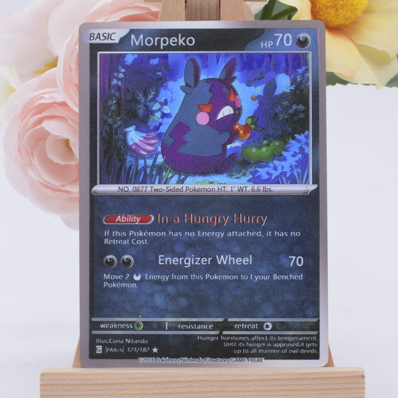 A single Pokémon trading card for Morpeko, displayed upright on a small wooden stand with soft pink and yellow flowers blurred in the background.