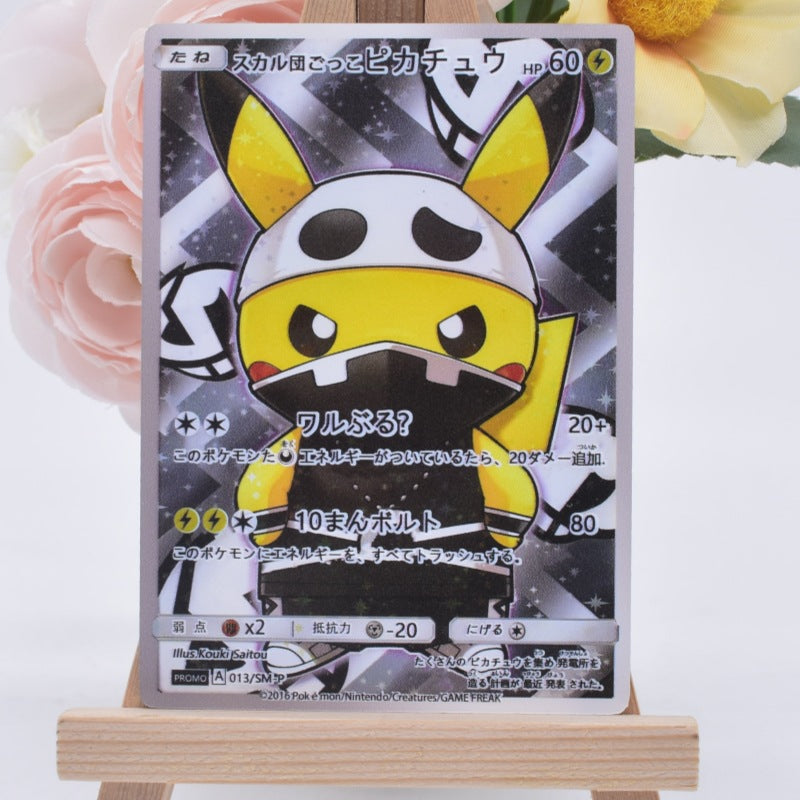A Pokémon trading card featuring a yellow and white character with black markings, displayed upright on a small wooden easel against a soft white background with blurred pink and yellow flowers.