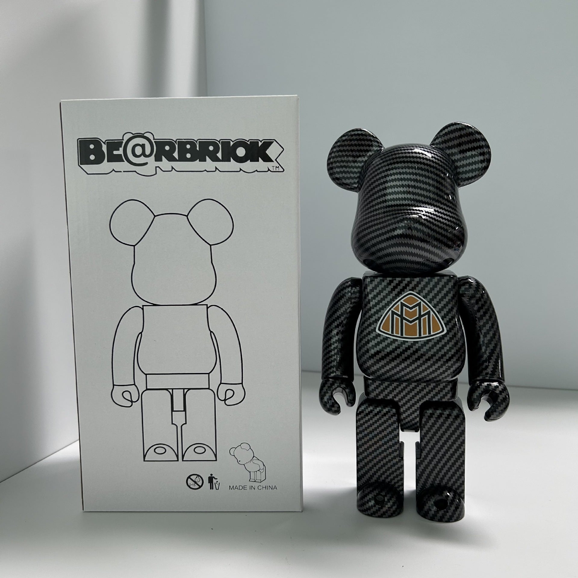 A Bearbrick collectible figure with a glossy black carbon fiber pattern stands next to its white packaging box, which features the 'BE@RBRICK' logo and a simple bear outline.