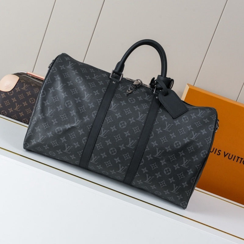 A Louis Vuitton monogram duffle bag in dark gray coated canvas with black leather handles and trim, displayed on a white shelf next to an orange Louis Vuitton shopping bag.